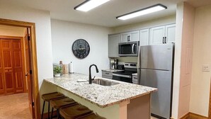 A modern kitchen with a central island, bar stools, stainless steel appliances, and a wall clock. - Targhee Creekside Condo - direct shuttle service to Targhee! (Driggs)