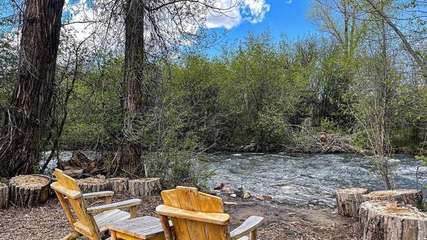 A wooden deck with two chairs and a table, surrounded by tree stumps and a river view. - Targhee Creekside Condo - direct shuttle service to Targhee! (Driggs)