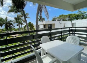 A balcony with white plastic chairs and a table, overlooking a lush green landscape with palm trees and a building in the background.