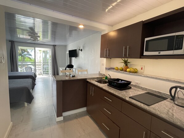 A modern kitchen with a microwave, a sink, and a countertop with a fruit bowl. There is a dining area with a table and chairs visible through a glass door.