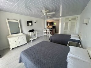 A bedroom with two beds, a dresser, a mirror, a ceiling fan, and a kitchenette in the background.