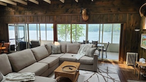 Interior acogedor de una cabaña con un amplio sofá modular, una mesa de centro de mimbre y vistas a un lago a través de amplios ventanales.