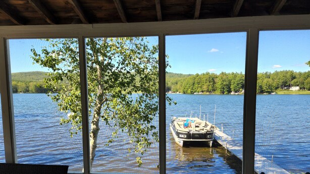 Una habitación con un gran ventanal con vista a un lago, un muelle con un bote y un árbol en primer plano.