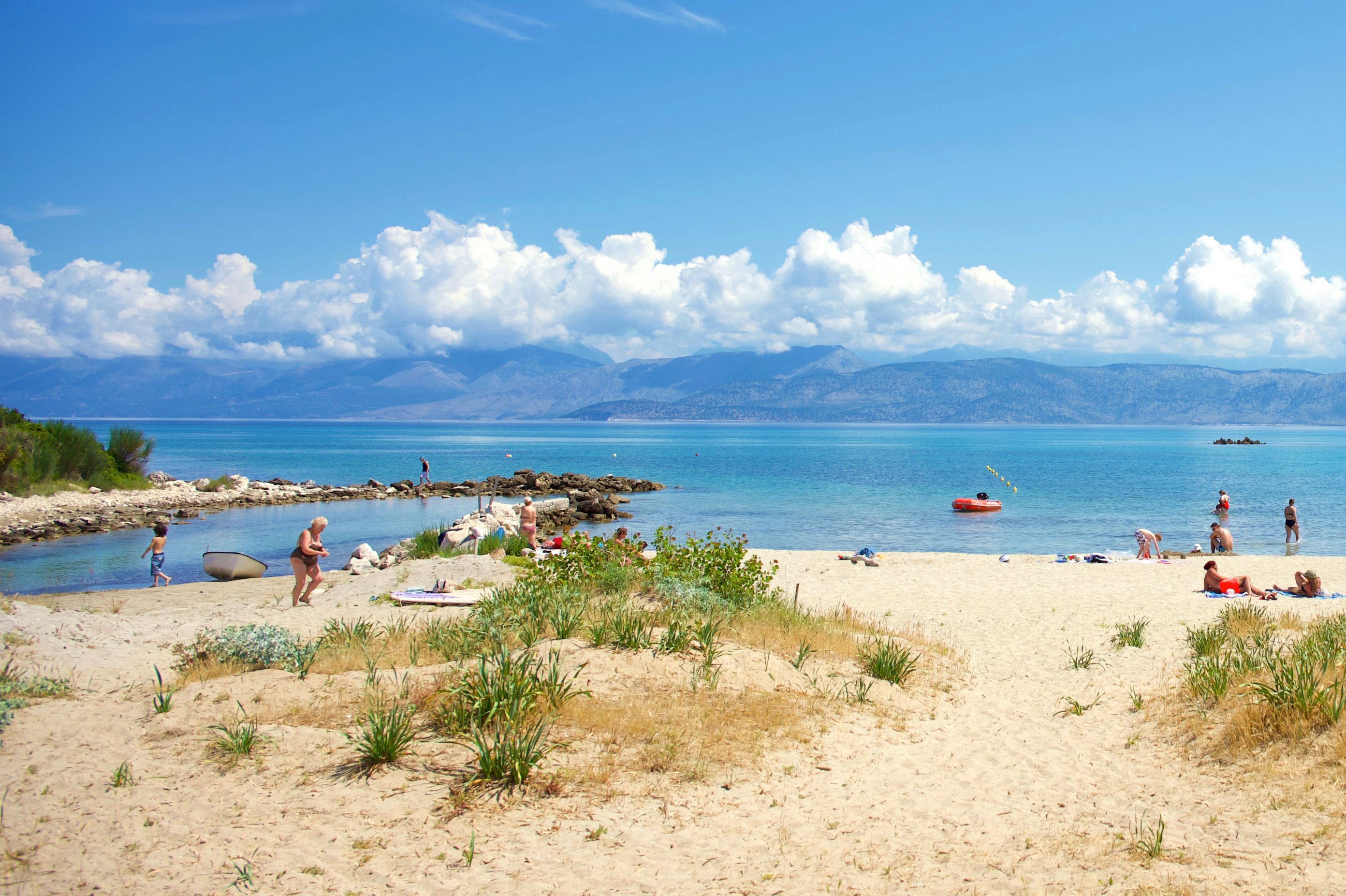 A beach with people sunbathing and swimming, rocky shoreline, and mountains in the background.