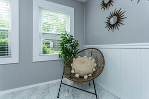 A room with a grey wall, a white-framed window, a wicker chair with a round cushion, and a potted plant.