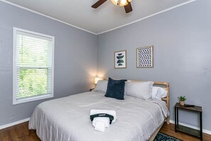 A bedroom with a bed, a window, a wall-mounted light, a small table, and a ceiling fan.