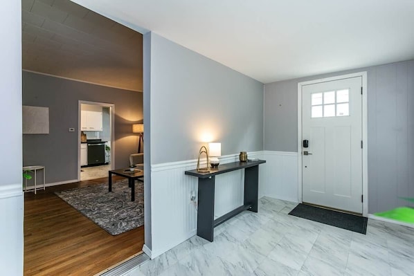A modern interior with a marble floor, a dark console table, a white door, and a kitchen visible in the background.