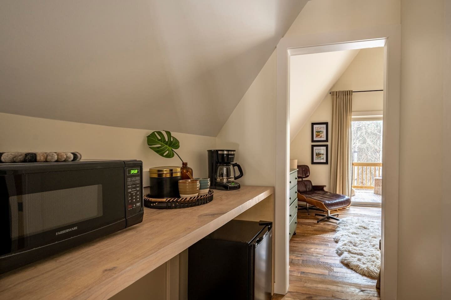 A compact kitchen with a microwave, coffee maker, and a small refrigerator. A wooden shelf holds decorative items, and a plant adds a touch of greenery. The kitchen leads to a balcony with a view of a snowy landscape.