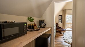 Une cuisine compacte équipée d’un four à micro-ondes, d’une machine à café et d’un petit réfrigérateur. Une étagère en bois expose des objets décoratifs, et une plante apporte une touche de verdure. La cuisine donne sur un balcon qui offre une vue sur un paysage enneigé.