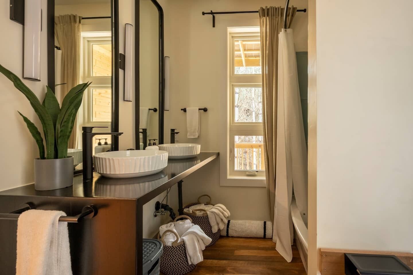 A bathroom with a large mirror, two sinks, a potted plant, and a window with curtains.