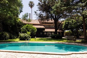 Une piscine dotée d’une terrasse en pierre, entourée d’une végétation luxuriante et bordée d’un bâtiment au toit recouvert de tuiles.