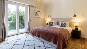 Ein Schlafzimmer mit einem großen Bett, zwei Nachttischlampen, einem Kronleuchter, Blick ins Grüne durch eine Schiebetür und einem gemusterten Teppich.