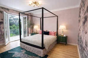 A four-poster bed with a checkered bedspread, two bedside tables with lamps, a green rug, and a view of greenery through a glass door.