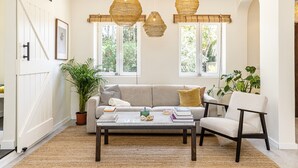 Ein gemütliches Wohnzimmer mit einer Couch, einem Sessel und einem Couchtisch, mit großen Fenstern und hängenden Korbtischen.
