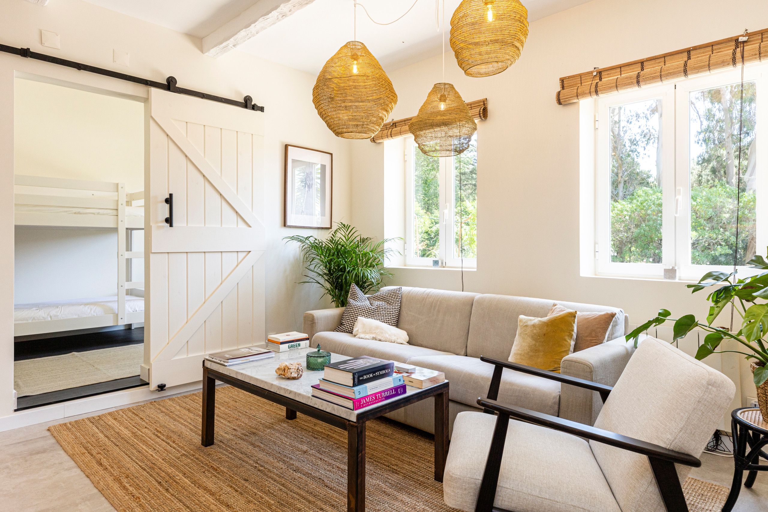 Un salon chaleureux avec un canapé, une table basse sur laquelle sont posés des livres, une plante et une vue sur l’extérieur grâce à de grandes fenêtres.
