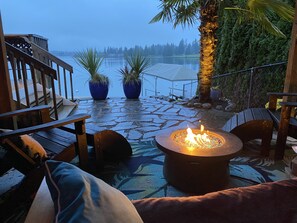A stone patio with a fire pit, outdoor seating, and a view of a lake. - Lakefront Studio Cabin & Tiki Hut. Enjoy Summer Fun and Winter Serenity!  (Lake Tapps)