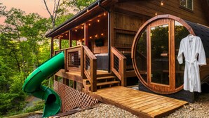 Une cabane en bois avec une terrasse en bois, un toboggan vert et un sauna.