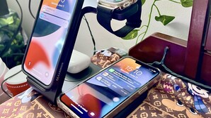 En smartphone på en holder sammen med en smartwatch, en bærbar computer og en Louis Vuitton taske på et bord.