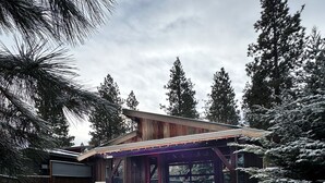 A wooden cabin with a covered patio and a grill, surrounded by snow-covered trees. - NW Modern, River, Old Mill near Downtown (Bend)