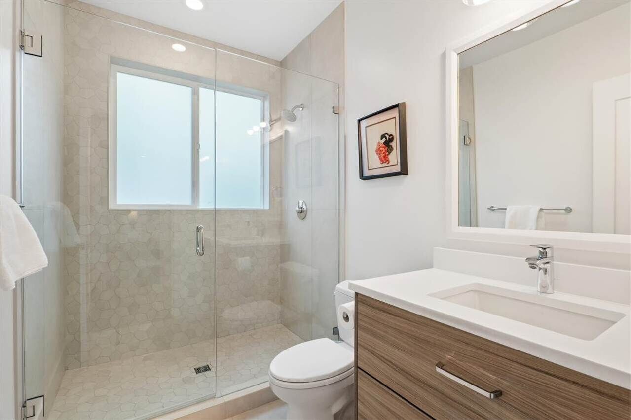 A modern bathroom with a glass-enclosed shower, a white toilet, and a wooden vanity with a large mirror.