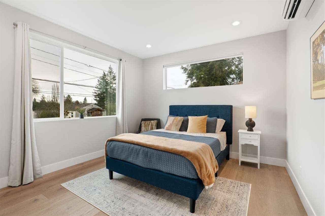 A bedroom with a bed, a nightstand, a window, and a view of trees and houses.