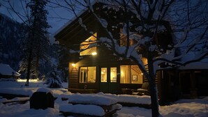 Una cabaña de madera con luz cálida en su interior, rodeada de nieve en el suelo y árboles.