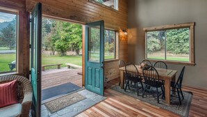 Interior de una cabaña de madera con comedor, una silla de mimbre y vistas a un paisaje verde.