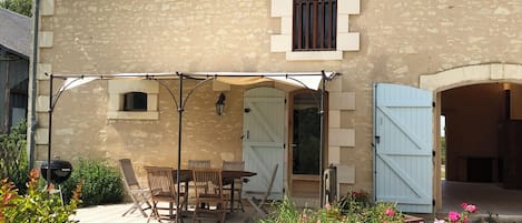 A stone building with a covered patio, a table with chairs, and a garden with flowers.