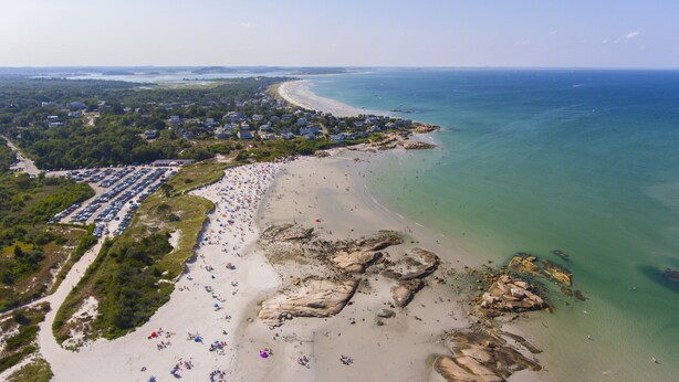 An aerial view of a beach with a parking lot, beachgoers, and a coastline.