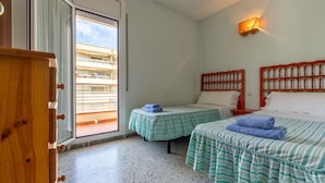 Habitación con dos camas, una mesita, un armario de madera y un balcón con vistas a edificios.