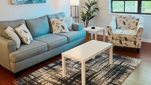 Un salón con un sofá gris, una mesa de centro blanca, un sillón estampado, una lámpara de pie y una ventana con vistas a la vegetación.