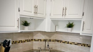 Una cocina con armarios blancos, encimeras de granito, un lavamanos doble y dos plantas en macetas sobre los estantes.
