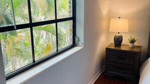 Una habitación con ventana que da a palmeras, una mesita de noche con lámpara y suelo de madera.