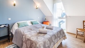 Una camera da letto con soffitto spiovente, un letto con copriletto decorato, due luci fissate al muro, un tavolino con un vaso e una sedia in legno.