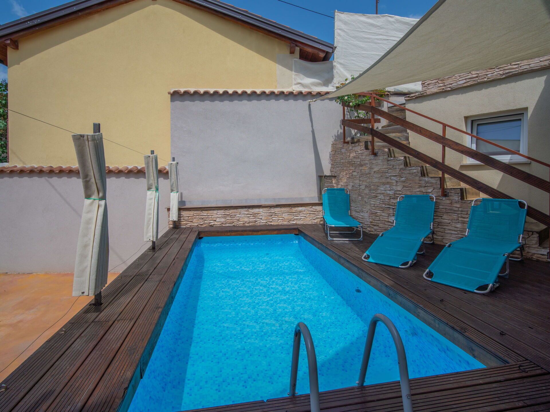 Et rektangulært swimmingpool med en træterrasse, to blå liggestole og en stenvæg.