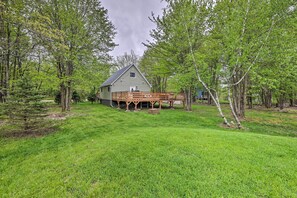 A rustic cabin with a wooden deck, surrounded by lush green trees and grass. - Near Lakes & Resorts: Pet-Friendly Long Pond Abode (Long Pond)