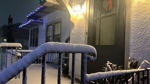 A snow-covered outdoor area with a metal railing, a building entrance with a lit wall, and a window with a red decoration. - Downtown Hibbing, 3 bd, 1 bath home with garage (Hibbing)