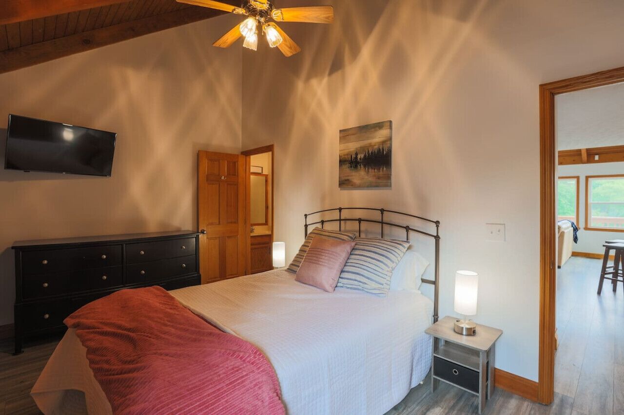 A bedroom with a bed, bedside table, TV, wooden dresser, and a door leading to another room.