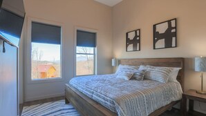 Une chambre à coucher avec un lit en bois, deux fenêtres équipées de stores, un tapis et deux œuvres d’art accrochées au mur.