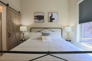 Un lit bien fait, recouvert d’une couvre-lit à motifs, deux tableaux accrochés au mur, une armoire en bois et une fenêtre avec des stores.