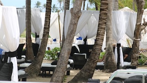 Un área de descanso frente a la playa con carpas blancas, sillas de salón y suelo arenoso.