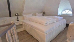 A bedroom with a large bed, white bedding, a nightstand with a lamp, and a window with a star decoration. - House Stedum Apartment 19 (Sylt)
