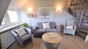 A modern living room with a grey sofa, two armchairs, a round wooden coffee table, and a spiral staircase. - House Stedum Apartment 19 (Sylt)