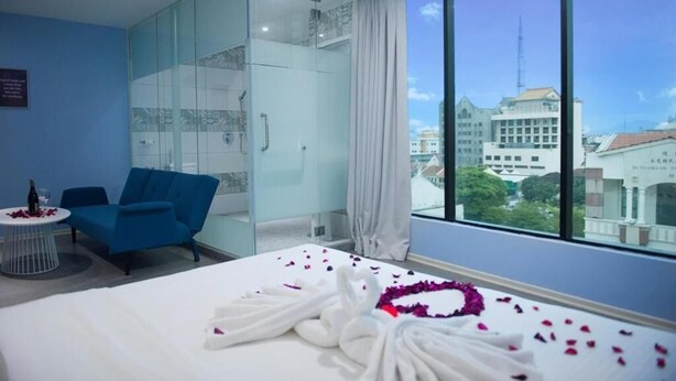 A hotel room with a bed, a blue armchair, a glass-enclosed wardrobe, and a large window offering a city view. - Q Collection @ Macalister(formerly know as Apple Hotel Penang)  (George Town)