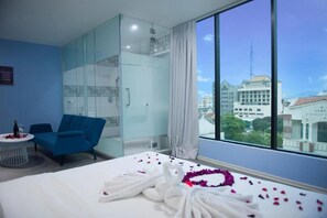 A hotel room with a bed, a blue armchair, a glass-enclosed wardrobe, and a large window offering a city view. - Q Collection @ Macalister(formerly know as Apple Hotel Penang)  (George Town)