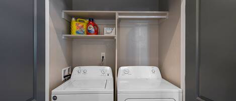 A laundry room with two white front-loading washers and dryers, shelves above containing cleaning supplies, and a power outlet on the left wall.