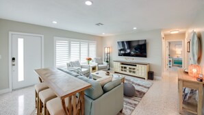 A living room with a sofa, coffee table, and TV. There is a dining table with chairs. The room has a door, windows with blinds, and a carpet.