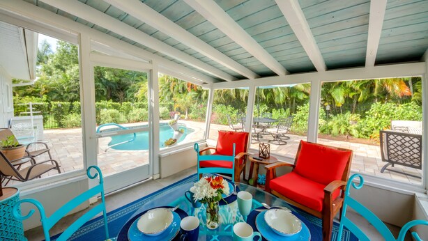 A covered outdoor seating area with a pool view, red and blue chairs, and a table with blue and white plates and cups.