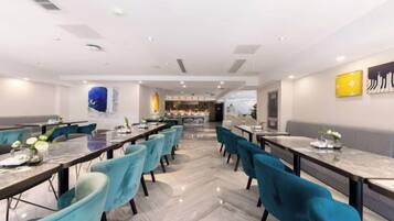 Un comedor moderno con mesas largas y sillas color turquesa, una cocina al fondo y vistas a la ciudad.