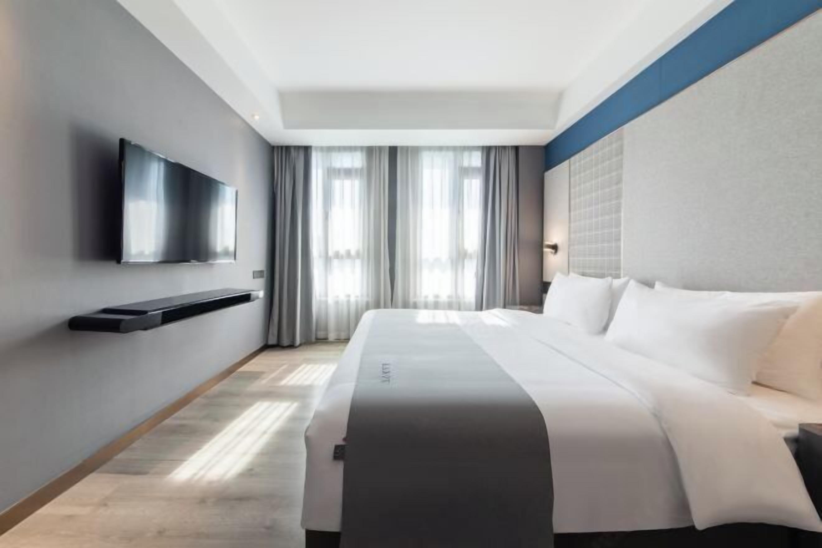 A modern hotel room with a large bed, a flat-screen TV mounted on the wall, and a window with curtains.